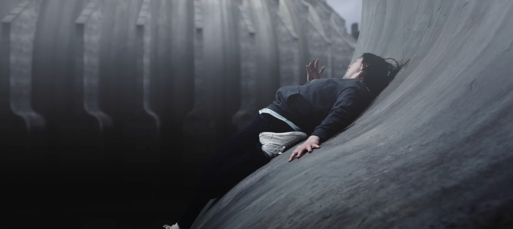 A still from short film 'Curve' (2021) by Tim Egan. Showing a woman on a slanting curved massive concrete structure precariously lying on the edge of a void.